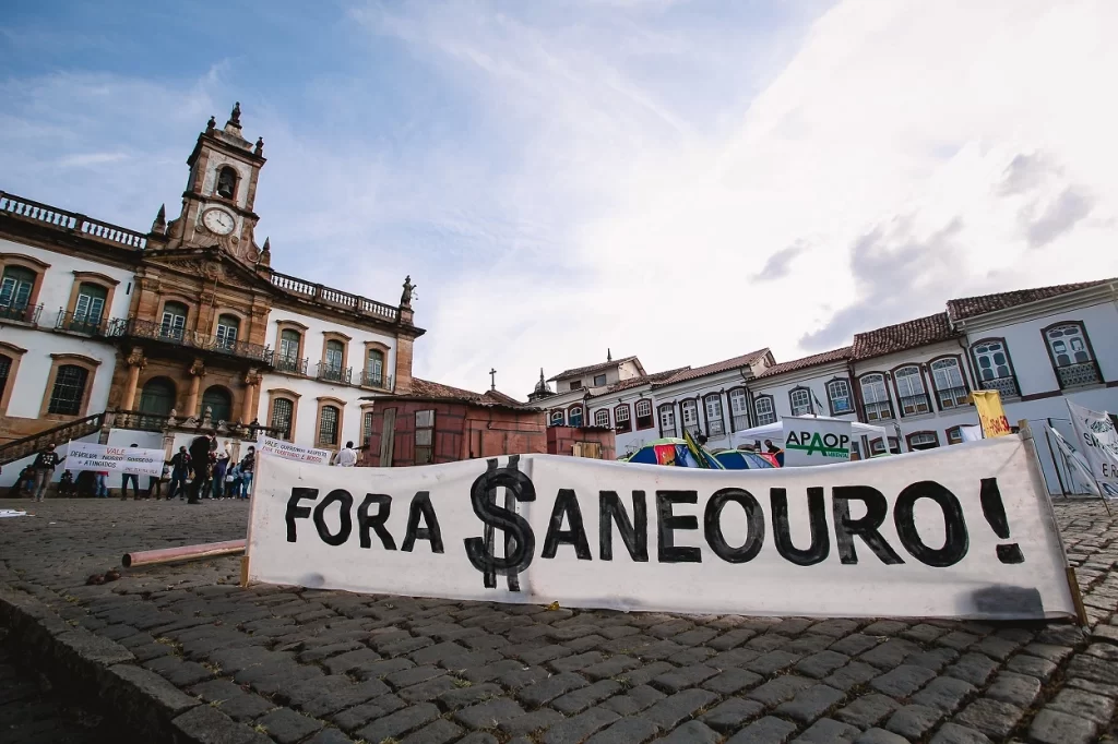 Praça Tiradentes, em Ouro Preto, ocupada por barracas do movimento Fora Saneouro., com o Museu da Inconfidência à esquerda e casas coloniais ao centro e à direita, ao fundo. À frente, a faixa com os dizeres “FORA SANEOURO”, com o símbolo do cifrão no lugar da letra S