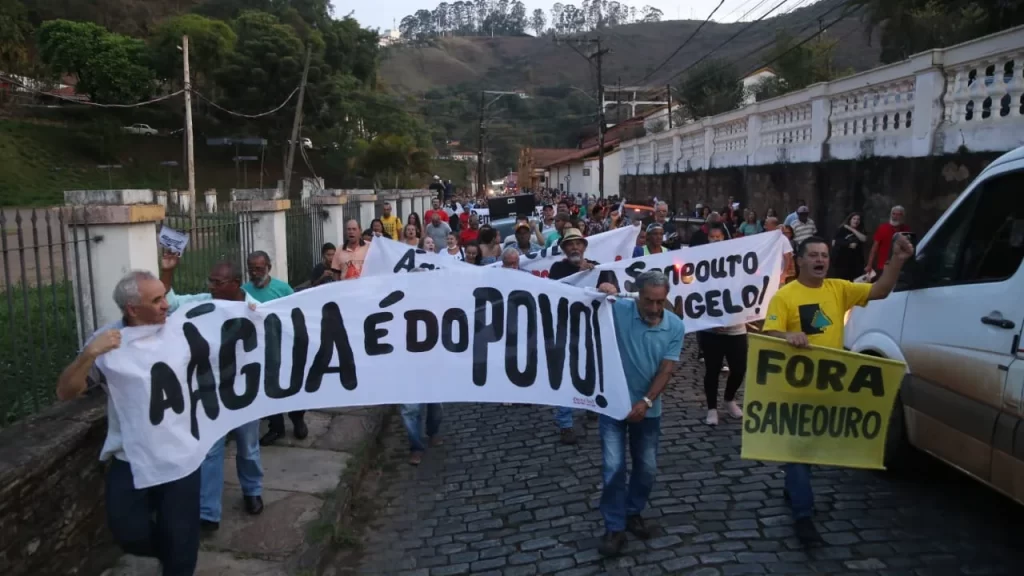 Pessoas participam de manifestação em Ouro Preto, portando faixas e cartazes com inscrições "A água é do povo" e "Fora Saneouro"