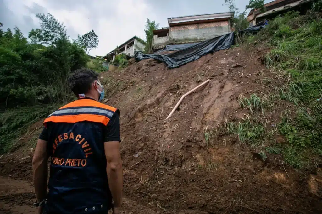 Homem, com colete preto e inscrição "Defesa Civil - Ouro Preto", observa encosta sob risco de desmoronamento durante o período chuvoso