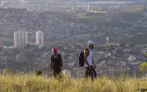 Dois jovens caminham em área de mata alta com vista panorâmica de cidade ao fundo, cena do curta que será exibido na Mostra Muito com Pouco.