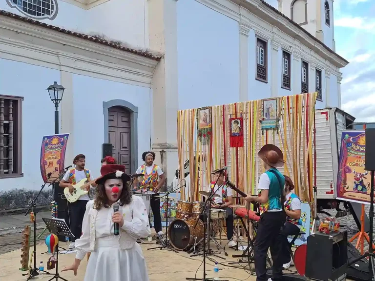 Banda com figurinos coloridos e palhaços se apresenta ao ar livre na Semana Mundial do Brincar, ao lado de uma igreja histórica.