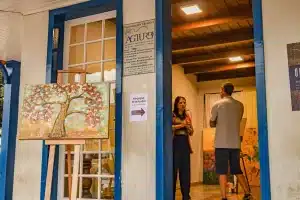 Na imagem se vê uma sala, com o artista e uma visitante. Também há um quadro ao lado de fora da porta e uma placa convidando as pessoas a visitarem a exposição de arte. Na sala acontecerá o projeto Ateliê Aberto.