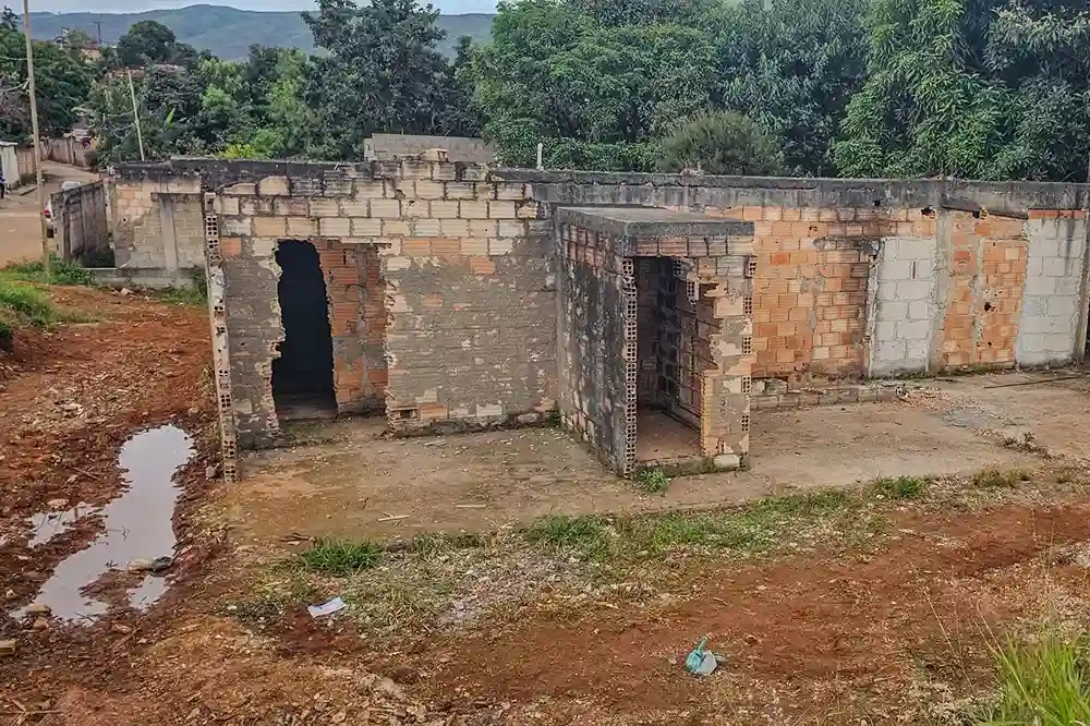 Estrutura abandonada de casa em ruínas. A construção está em um terreno de terra batida com poça d’água à frente, e há vegetação ao fundo.