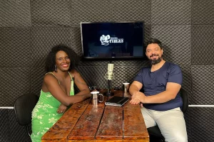 No estúdio da Agência Primaz, uma tv de fundo exibe a logo da Entrevista Primaz, no lado esquerdo da Mesa vemos Danielle dos Anjos, mulher negra de cabelo black power e vestido, do lado direito da mesa, o jornalista Lui Pereira. No centro da mesa tem um microfone condensador e alguns objetos como canecas e tablet