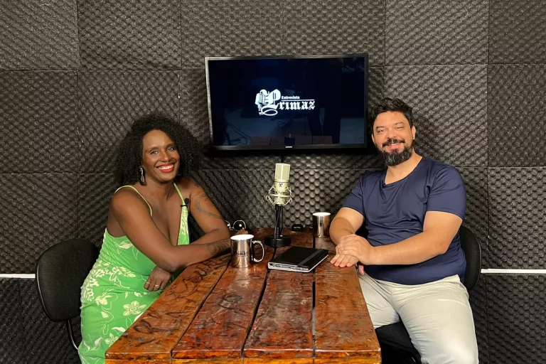 No estúdio da Agência Primaz, uma tv de fundo exibe a logo da Entrevista Primaz, no lado esquerdo da Mesa vemos Danielle dos Anjos, mulher negra de cabelo black power e vestido, do lado direito da mesa, o jornalista Lui Pereira. No centro da mesa tem um microfone condensador e alguns objetos como canecas e tablet