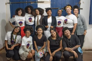 Diversas mulheres negras sorriem e posam para uma foto em grupo, todas elas olham para a câmera, elas estão dentro de uma casa e ao fundo tem duas janelas azuis de madeira