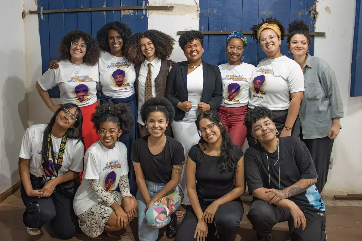 Diversas mulheres negras sorriem e posam para uma foto em grupo, todas elas olham para a câmera, elas estão dentro de uma casa e ao fundo tem duas janelas azuis de madeira