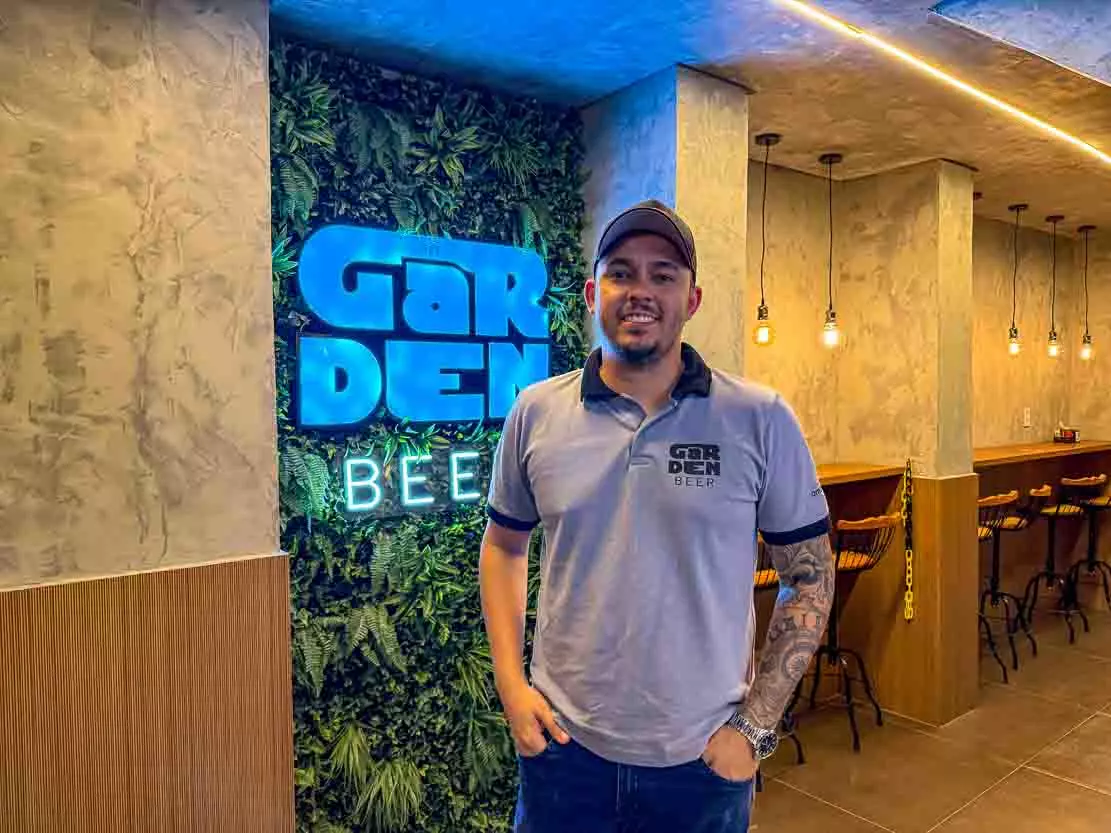 O proprietário do Garden Beer, Filipe Hilder posa de frente para uma placa, dentro do seu bar, com o nome do estabelecimento