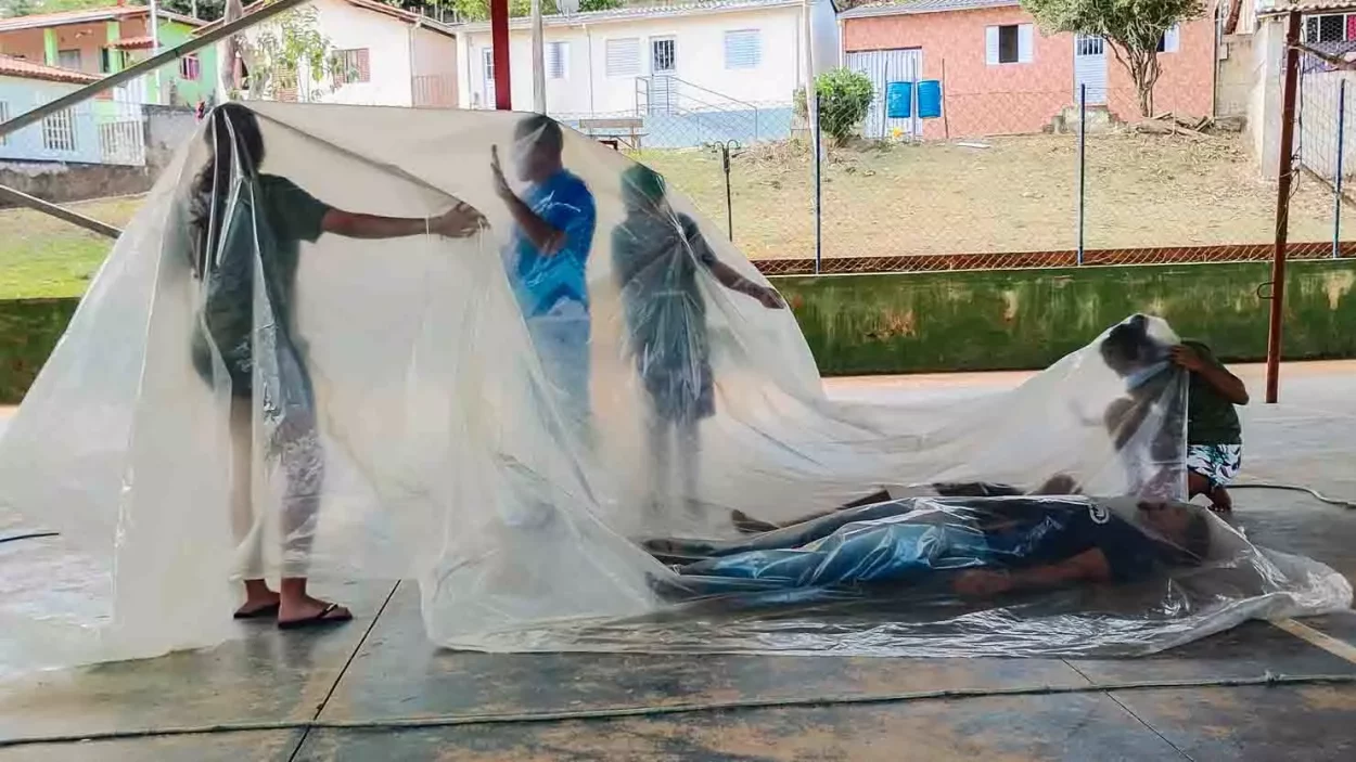 As pessoas estão embaixo de uma lona plástica semi transparente dentro da quadra de gesteira, elas realizam atividades teatrais