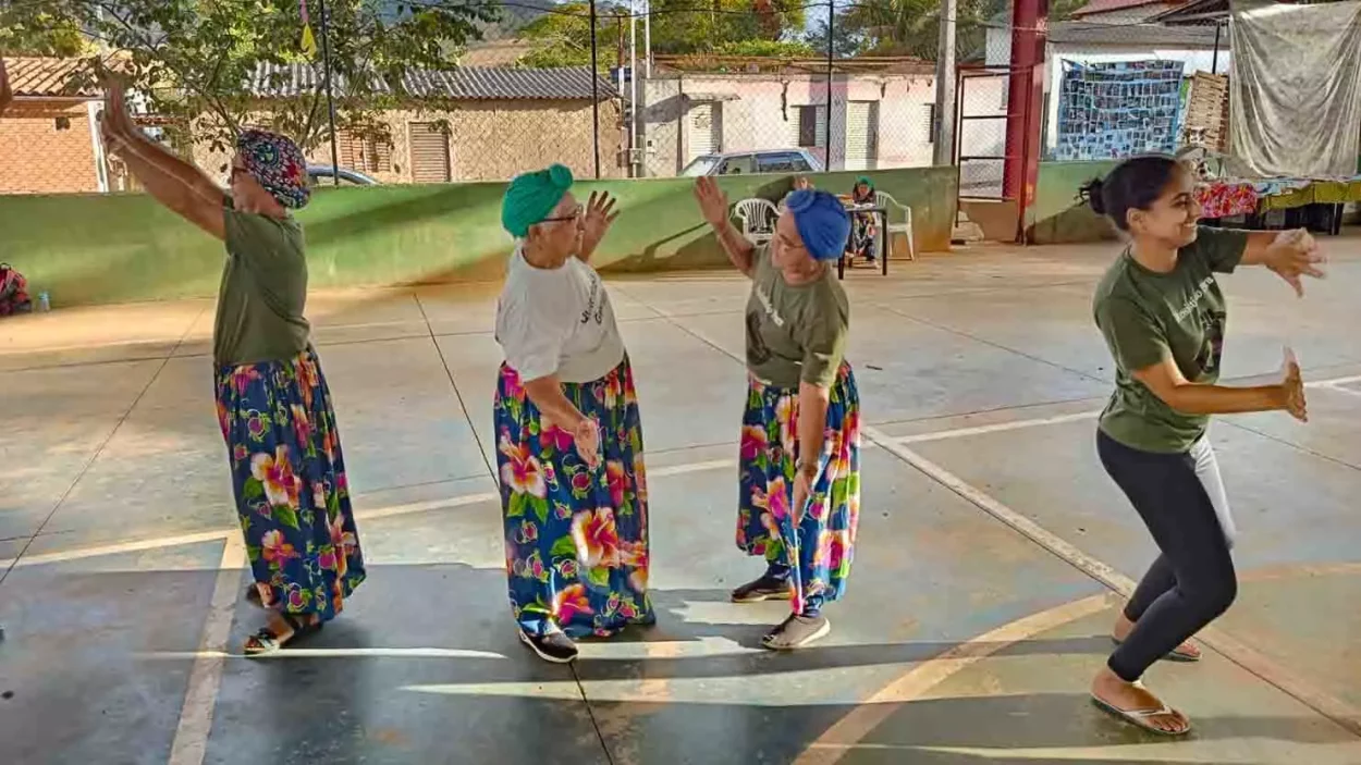 No centro da imagem, duas mulheres idosas com saias coloridas e blusas brancas se encaram e parecem copiar uma o movimento da outra, como um espelho. Atrás de cada uma delas, outras duas meninas parecem fazer o mesmo exercício com outras pessoas extra quadro