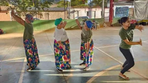 No centro da imagem, duas mulheres idosas com saias coloridas e blusas brancas se encaram e parecem copiar uma o movimento da outra, como um espelho. Atrás de cada uma delas, outras duas meninas parecem fazer o mesmo exercício com outras pessoas extra quadro