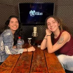 duas mulheres sorriem para a câmera, elas estão sentadas uma de frente para a outra, uma mesa as separa, ao fundo uma tv exibe a logo da Entrevista Primaz e sobre a mesa tem um tablet, um microfone e algumas canecas