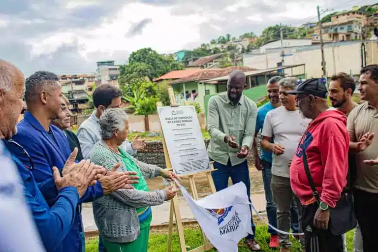 A imagem mostra prefeito, vice prefeita, vereadores e moradores dos bairros Barro Preto e Morada do sol descerrando a placa de inauguração da nova bacia de contenção de Mariana, ao fundo, vemos a bacia.