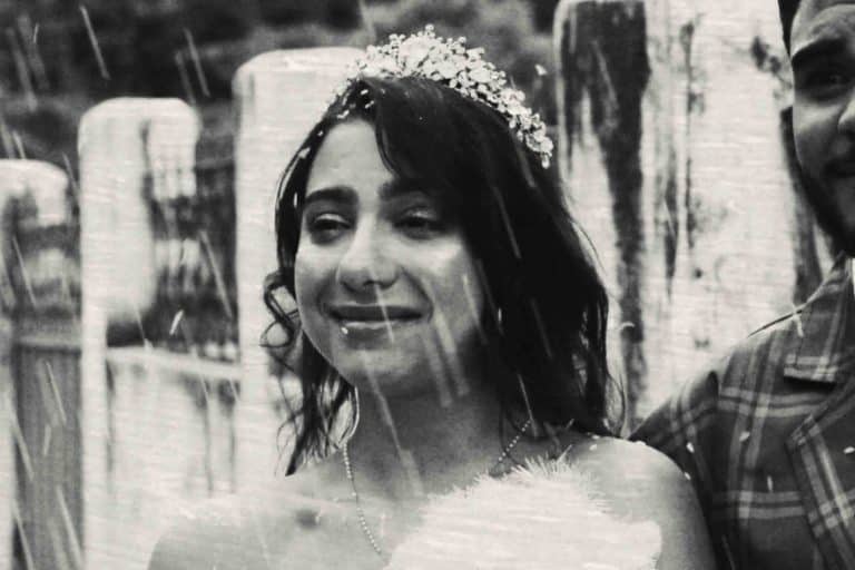 Fotografia em preto e branco, em plano médio, de uma mulher jovem com expressão serena e um leve sorriso. Ela tem cabelos escuros e ondulados, e usa uma tiara delicada com detalhes de pedrarias e flores sobre a cabeça. Ela veste o que parece ser um traje especial, com um colar fino no pescoço, e segura algo felpudo e branco à frente do corpo, possivelmente um buquê. O ambiente é externo e rústico, com colunas de pedra ao fundo. Fortes traços de chuva caem diagonalmente em frente à câmera, criando uma atmosfera cinematográfica e sensível. À direita, aparece parte do rosto e o ombro de um homem vestindo uma camisa xadrez, sugerindo que ela está acompanhada.