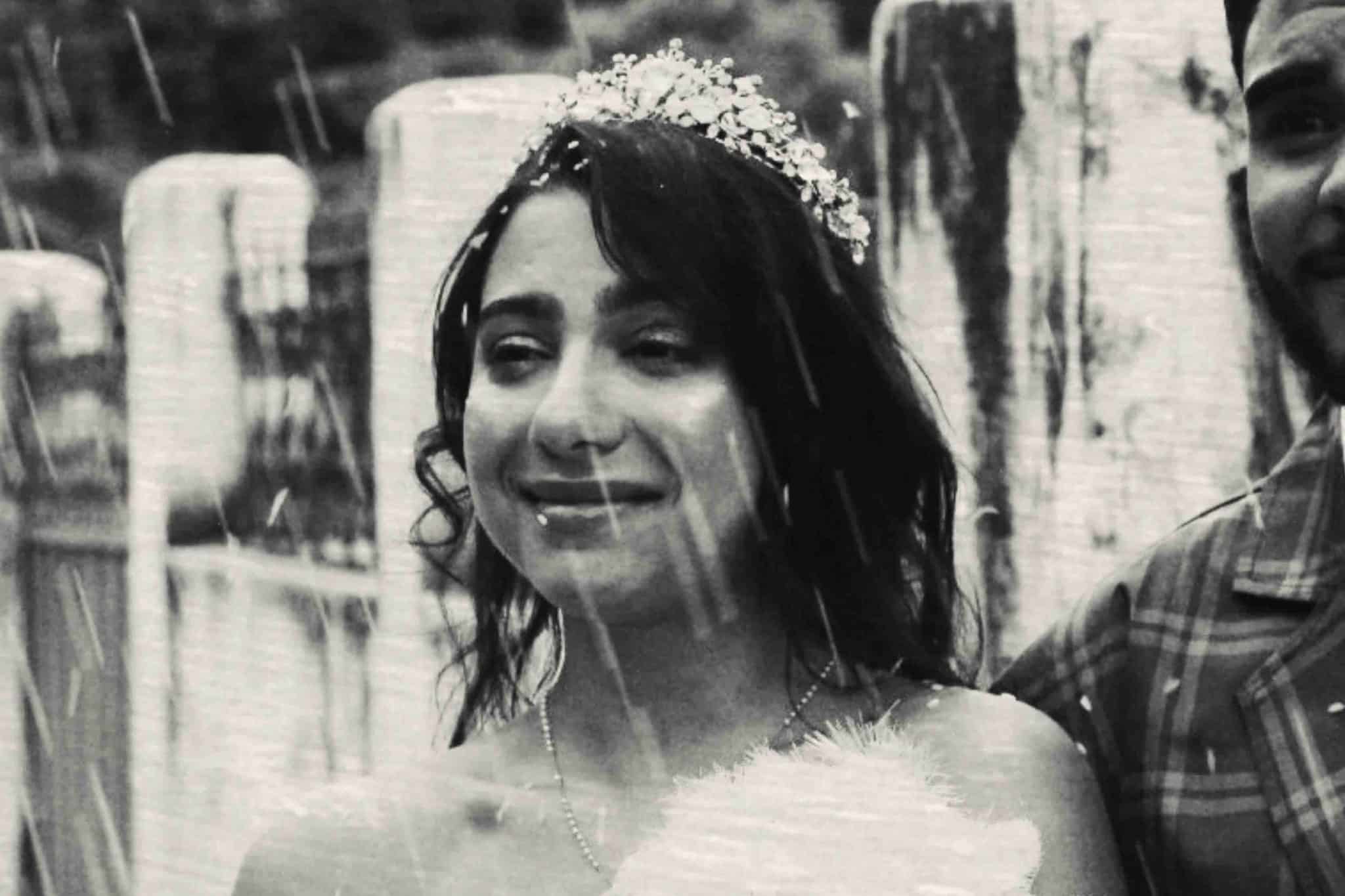 Fotografia em preto e branco, em plano médio, de uma mulher jovem com expressão serena e um leve sorriso. Ela tem cabelos escuros e ondulados, e usa uma tiara delicada com detalhes de pedrarias e flores sobre a cabeça. Ela veste o que parece ser um traje especial, com um colar fino no pescoço, e segura algo felpudo e branco à frente do corpo, possivelmente um buquê. O ambiente é externo e rústico, com colunas de pedra ao fundo. Fortes traços de chuva caem diagonalmente em frente à câmera, criando uma atmosfera cinematográfica e sensível. À direita, aparece parte do rosto e o ombro de um homem vestindo uma camisa xadrez, sugerindo que ela está acompanhada.