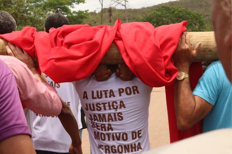 A foto, tirada de costas em um ângulo médio, mostra um grupo de pessoas carregando coletivamente um grande tronco de madeira sobre os ombros. O tronco está parcialmente coberto por um tecido vermelho vibrante que cai pelas laterais. No centro da imagem, um homem veste uma camiseta branca com uma frase de protesto escrita em letras maiúsculas de cor marrom: "A LUTA POR JUSTIÇA JAMAIS SERÁ MOTIVO DE VERGONHA" Detalhes da cena: Composição: Outras pessoas aparecem nas bordas da foto, ajudando a sustentar o peso do tronco. À direita, vê-se o braço de um homem com um relógio dourado e uma camiseta azul; à esquerda, parte de uma camiseta rosa e outra branca. Ambiente: O cenário ao fundo é ao ar livre, mostrando uma estrada de terra ou asfalto claro, cercada por árvores verdes e morros sob um céu nublado. Atmosfera: A imagem transmite um sentimento de esforço coletivo, resistência e mobilização social.