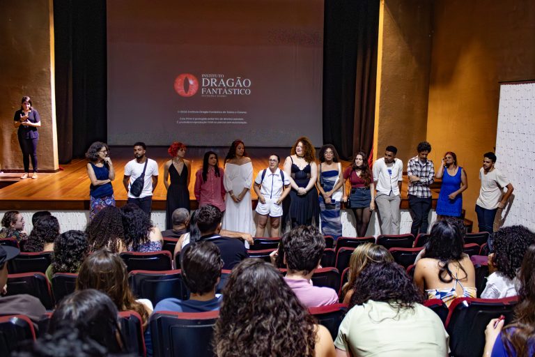 Uma foto horizontal, de plano médio, mostra um grupo diverso de treze pessoas, homens e mulheres, em pé em um palco de teatro, de frente para uma plateia lotada. Eles estão alinhados em frente a uma grande tela de projeção que exibe o logotipo vermelho e branco do "Instituto Dragão Fantástico" e texto de direitos autorais. Uma mulher na extrema esquerda do palco segura um microfone e gesticula enquanto fala. As pessoas no palco usam uma variedade de roupas casuais e formais. A plateia, vista das fileiras de trás, preenche o teatro e está voltada para o palco, com as costas das cabeças visíveis. O teatro tem paredes escuras e cortinas pretas ao lado da tela. A iluminação é focada no palco.