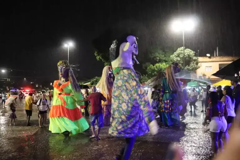 A imagem mostra os catitões (bonecos gigantes feitos de papel machê e pano) dançando à noite, embaixo de chuva na Praça dos Ferroviários, em Mariana